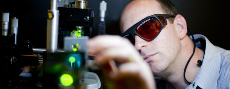 Engineer wearing safety glasses carefully adjusting a lab instrument that is emitting a green laser beam.